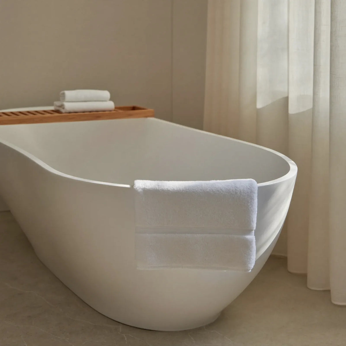 A tidy bathroom in natural light, with a white bath, pale walls and towels folded on a wooden shelf