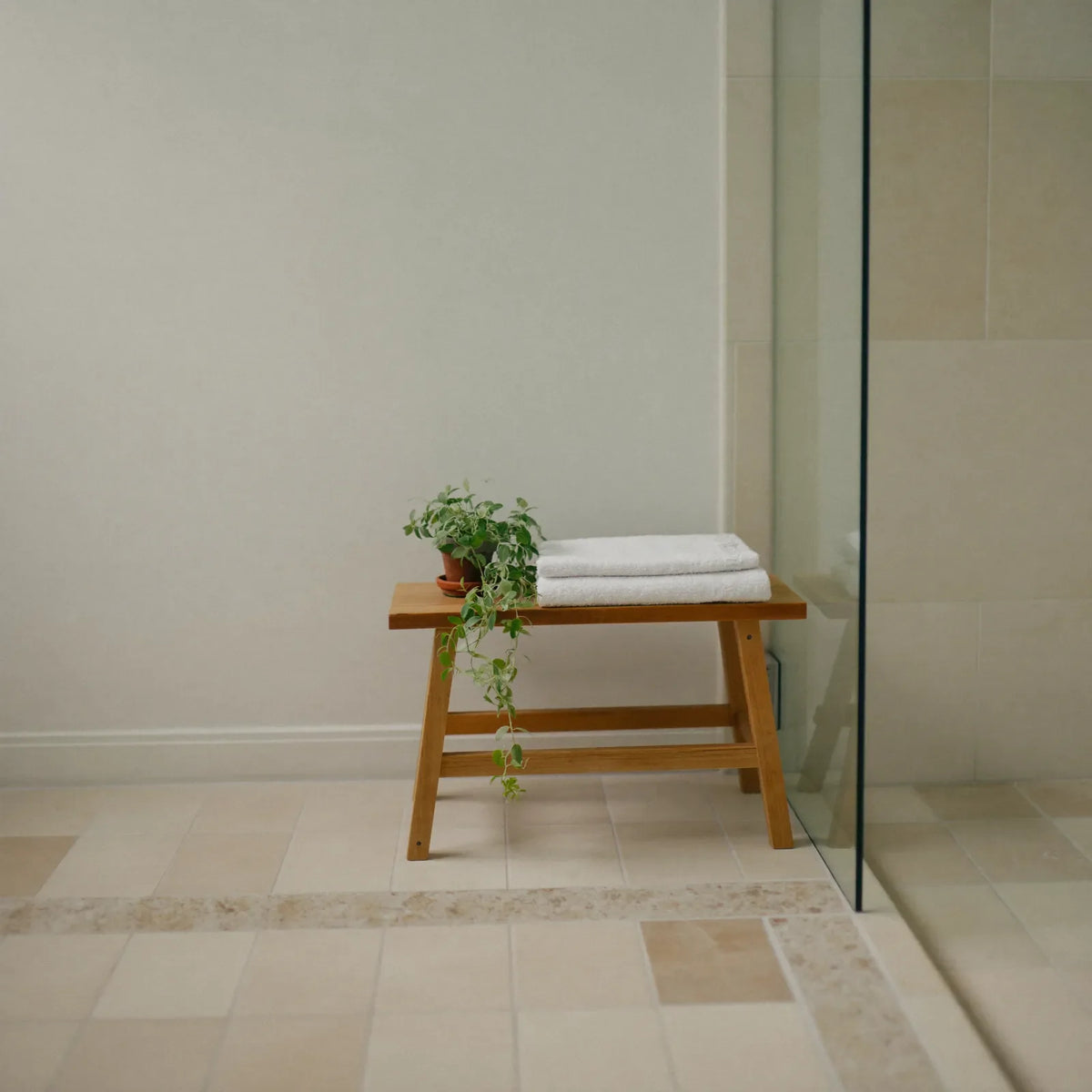 A quiet bathroom in early morning light, with a white bath, pale tiles and a folded towel on a wooden stool