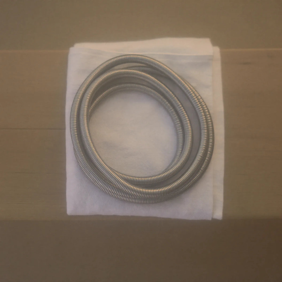 A coiled silver metal shower hose resting on a folded white cloth on a pale wooden surface