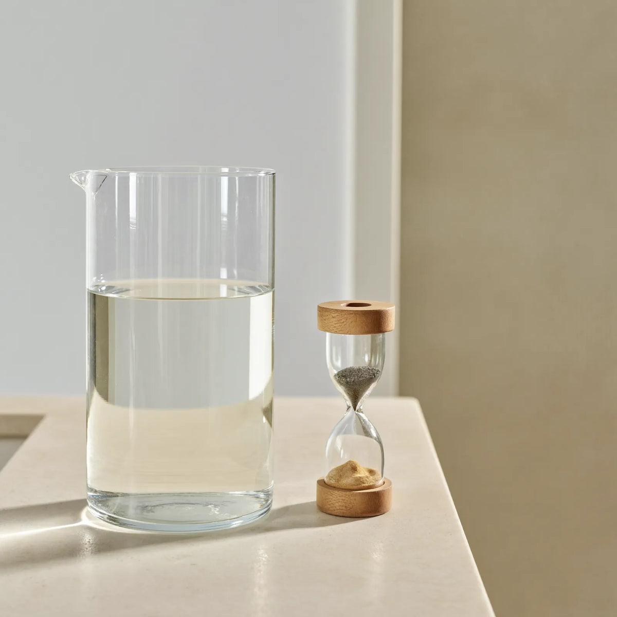 A clear glass jug of water beside a small wooden sand timer on a pale stone worktop