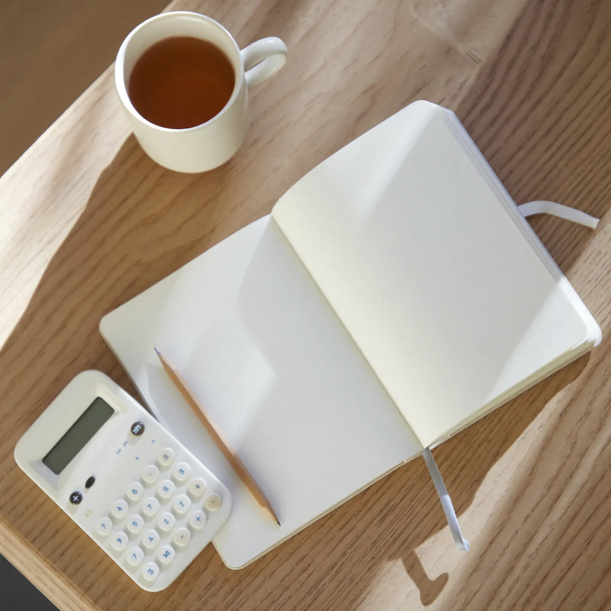 An open notebook with a pencil, a white calculator and a mug of tea on a light wooden table
