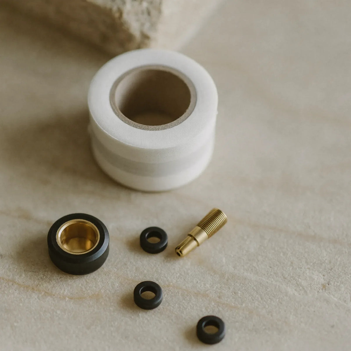 A roll of white PTFE tape, a brass-lined hose nut, a small threaded brass fitting and three black washers on pale stone