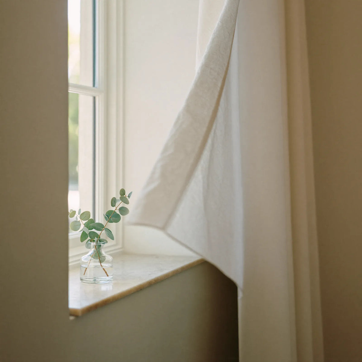A small glass vase of eucalyptus on a sunlit window sill beside a white curtain lifting in the breeze